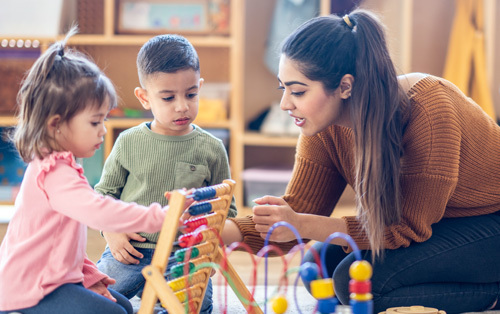 Female preschool teacher sits on the floor with students as they play with various toys 